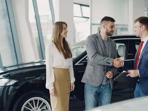 Customers shaking hands with a JE Auto salesperson beside a dark saloon